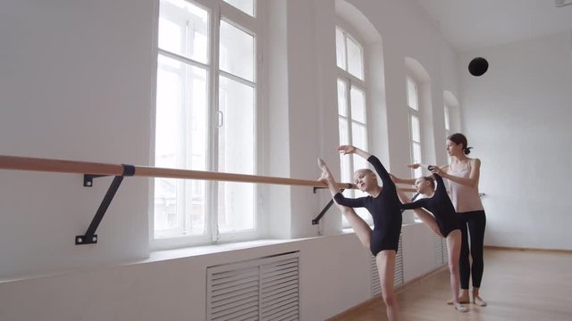 Female ballet teacher helping two little Caucasian girls in black costumes standing with their legs on barre and stretching