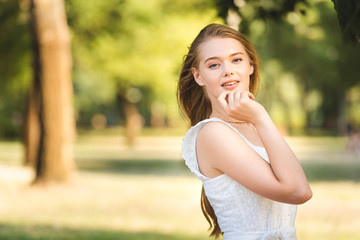 front view of beautiful young girl in white dress posing while standing in park and looking at camera