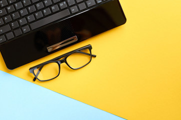 Black glasses and laptop keyboard on blue and yellow background composition.