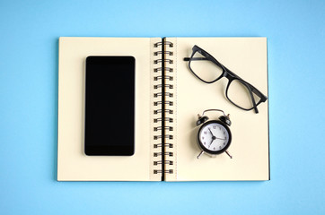 Black glasses, alarm clock, paper notepad and cellphone on blue background composition.