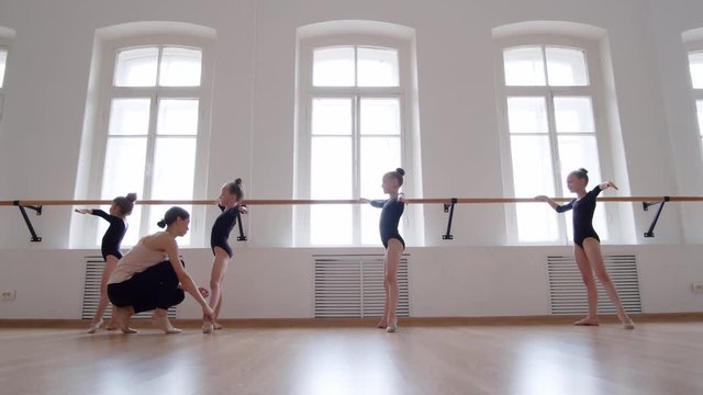 Wide shot of young Caucasian woman going along dance studio and explaining little ballerinas standing in right position at barre