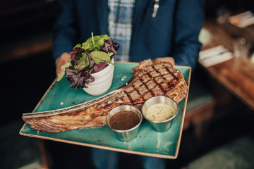 A restaurant waiter is holding a plate with Tomahawk steak served with salad and sauces. Concept of...