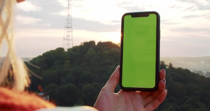 Close-up of girl using smartphone taking picturs of sunset and landscape. Female hand tapping touching mock-up green chromakey screen browsing in app outdoors.