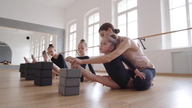 Pan shot of three little girls in black costumes sitting on floor in split using yoga blocks and watching female ballet coach helping their teammate stretching her legs