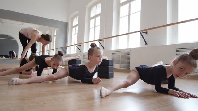 Zoom out of four little female dancers in black leotards sitting on floor in twine using yoga blocks while young female ballet teacher helping one of them