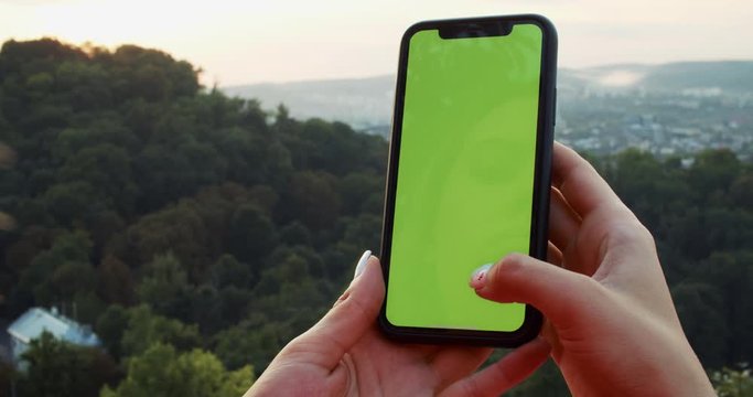 Close-up young woman holding mock-up smartphone browsing tapping on modern chromekey greenscreen staying in stunning nature landscape. Urban cityscape in background.