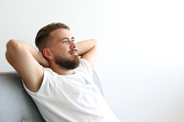 Bearded guy wearing blank white t-shirt and denim pants sitting alone at home on grey textile couch. Young man with facial hair in domestic situations. Interior background, copy space, close up.