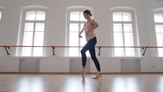 Wide shot of young ballerina training in studio while listening to music through wireless earphones and using her smartphone