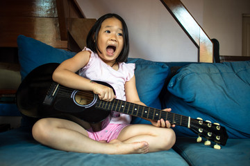 Asian playful girl sitting on blue sofa at home to sing a song with guitar