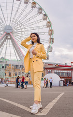 Young and stylish girl walks around the city opposite the ferris wheel in yellow clasic suit. Fashion.