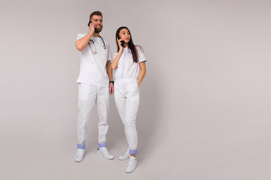 Two Stylish Nurses, Male And Female, In White Casual Costumes Are Standing Like Team And Talking On The Mobile Phones On A White Wall Background. Medical Lifestyle Concept. Free Space On Right Side