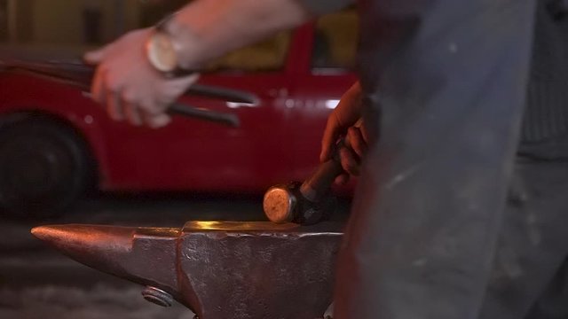 Medium close-up shot of a knifemaker forging a hot orange glowing knife with a big hammer on an old anvil outside in the dark night - Slow Motion