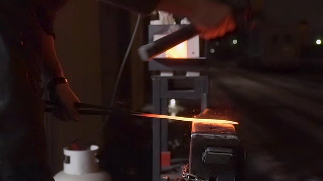 Medium side shot of hard-working knifemaker forging a hot orange glowing knife with a big hammer on an old anvil outside, in the dark night, with burning forge in background - Slow Motion