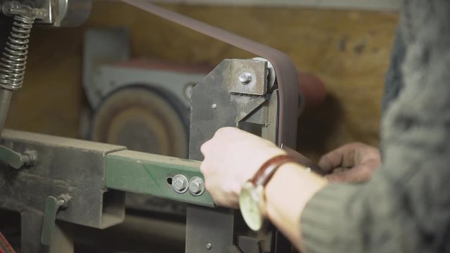 Medium close-up shot of a knifemaker sharpening a knife on a belt sander bench in his workshop - Slow Motion