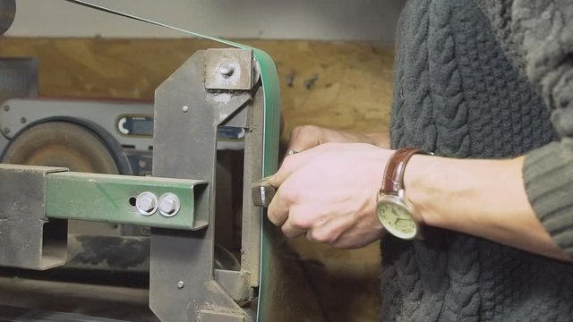 Medium close-up shot of knifemaker with mask sanding a kitchen knife's wood handle on a belt sander bench in his workshop - Slow Motion