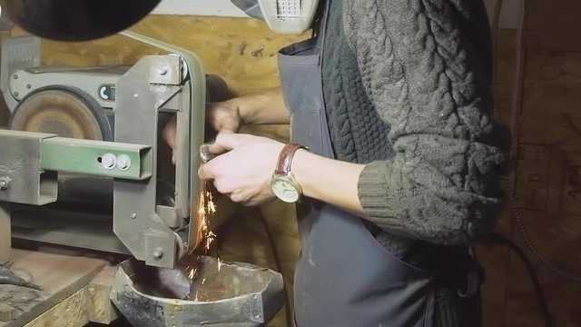 Medium shot of knifemaker with mask making sparks while sanding a kitchen knife's wood handle on a belt sander bench in his workshop - Slow Motion