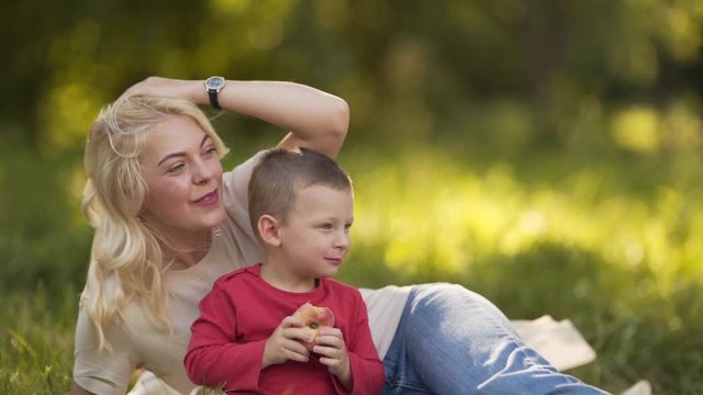 A blond woman lying in the park, hugging and kissing her kid as he is eating a donut