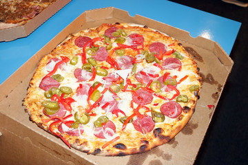 A cheap pizza with salami in a cardboard box lies on the edge of the table