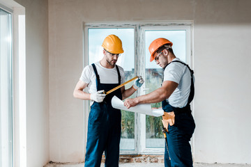 handsome builder looking at blueprint near coworker holding measuring tape
