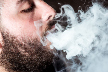 Detail of a young man with pigtail exhales smoke through the nose of the electronic cigarette on black background