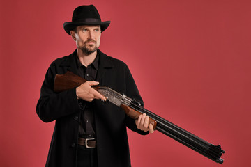 Middle-aged man with beard, mustache, in black jacket and hat, holding a gun while posing against a red background. Sincere emotions concept.