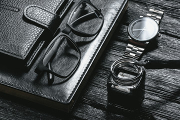 Business concept background. Black leather book, inkwell with a quill pen, wallet, eye glasses and a hand watch on a black wooden table.