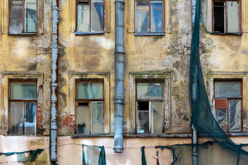 Fototapeta na wymiar An old residential building awaiting reconstruction or demolition. Facade with windows of crumbling plaster, scraps of green protective mesh and rusty drainpipes