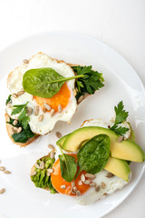 Fried eggs on a toasts bread with avocado, spinach and seeds on a white plate on the white background.