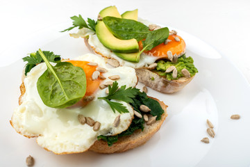 Fried eggs on a toasts bread with avocado, spinach and seeds on a white plate on the white background.