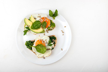 Fried eggs on a toasts bread with avocado, spinach and seeds on a white plate on the white background.