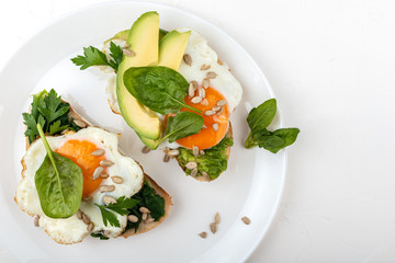 Fried eggs on a toasts bread with avocado, spinach and seeds on a white plate on the white background.