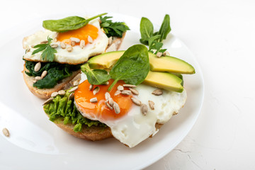 Fried eggs on a toasts bread with avocado, spinach and seeds on a white plate on the white background.