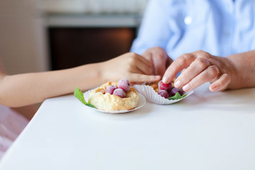 Senior woman and child girl with homemade pastries at kitchen. Hands of kid and retired grandmother. Baked buns with berries and mint leaves. Sweets with powdered sugar.