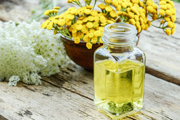 Glass bottle of essential oil or tincture of extract near yellow fresh flowers of tansy and yarrow on wooden background. Herbal medicine concept.