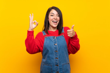Young Mexican woman with overalls over yellow wall showing ok sign and thumb up gesture