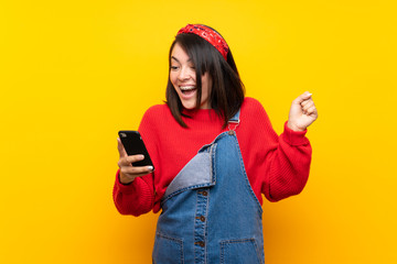 Young Mexican woman with overalls over yellow wall surprised and sending a message