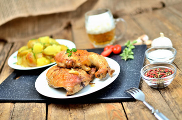 Golden grilled chicken legs on a plate, roasted potatoes and cold beer, rosemary, tomatoes, oregano, salt, pepper and garlic on a wooden table
