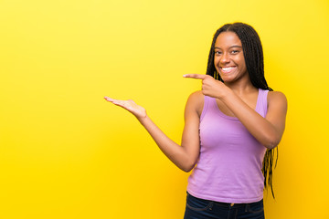 African American teenager girl with long braided hair over isolated yellow wall holding copyspace imaginary on the palm to insert an ad