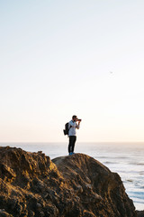 Hipster young man hiker standing on cliffs photographing sunset over ocean