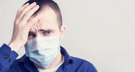 Young man in a medical mask, he has a headache, squeezes his head with his hands, sad looking at the camera, close up, toned