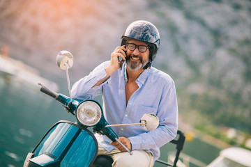 Mature man on motor scooter using phone in marina.