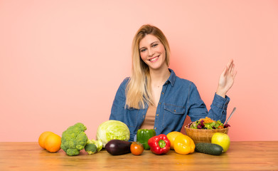 Young blonde woman with many vegetables saluting with hand with happy expression