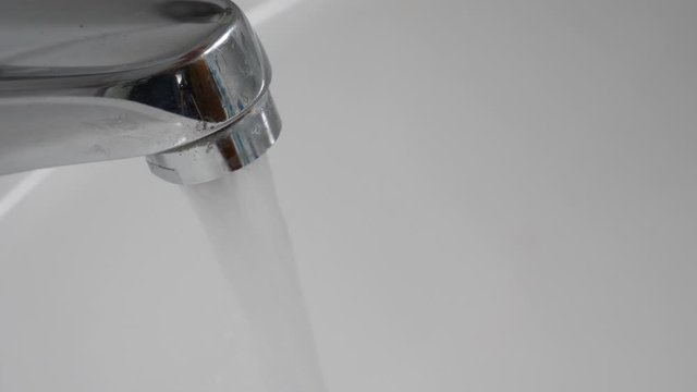 Tramsparent water running from chromium plated or metal tap into white sink in bathroom. Slow motion close-up shot. No people. Continious off on.