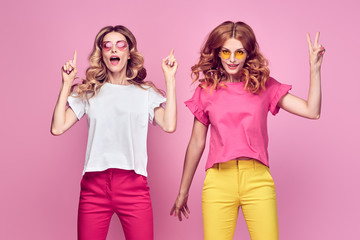 Obraz na płótnie Canvas Two fashionable girl jump Smiling in colorful outfit on pink. Beautiful easy-going woman in red yellow pants, Stylish curly hair having fun. Joyful funny slim sisters friends, happy fashion concept