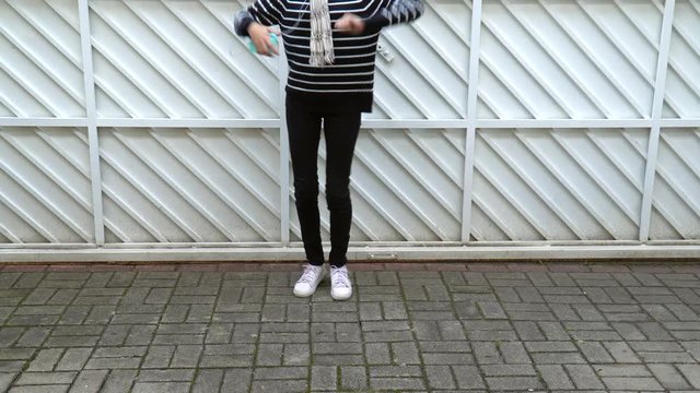 Young girl dancing and listening music on cellphone on white gate background. Unrecognizable model body holding smartphone and earphones wearing casual black pants and pullover and white sneakers.