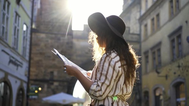 Standing young woman tourist on the street of old town Prague at morning time. Medium shot attractive girl looking at map, spinning and glance around in backlit, she visiting the sights in holiday