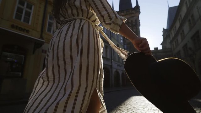 Following from the back woman tourist walking in old city centre of Prague at morning time. Low angle view unrecognizable female in backlit goes down street and flapping brown hat in hand