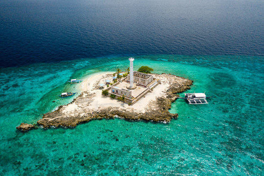 Aerial drone view of traditional boats next to a tiny tropical island with a lighthouse, surrounded by coral reef (Capitancillo Island, Philippines)