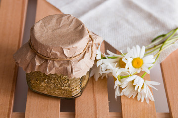 a jar of herbal tea stands on a wooden tray of rekk with a daisy flower and a beige coarse cloth...