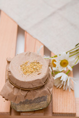 a jar of herbal tea stands on a wooden tray of rekk with a daisy flower and a beige coarse cloth copy space top view